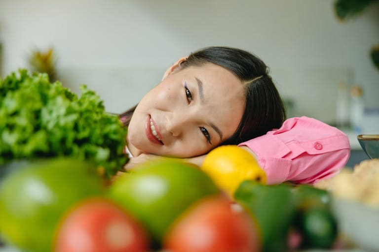 Young woman in pink shirt posing with fresh vegetables and fruits, smiling indoors.