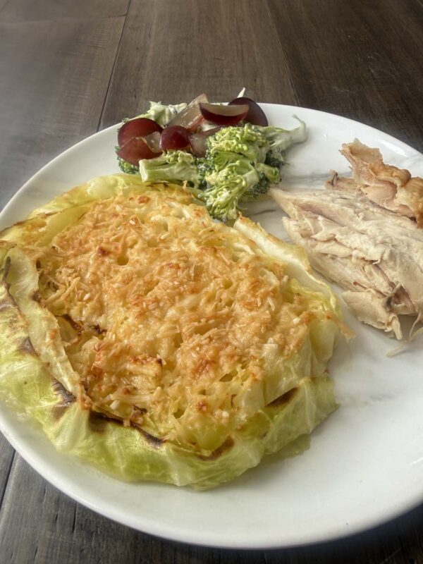 a cheesy cabbage steak, broccoli and grape salad, and chicken on a white plate