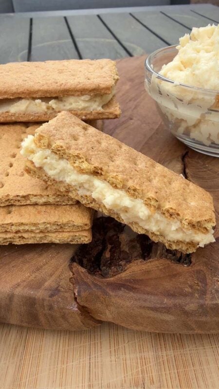 graham cracker sandwiches filled with protein frosting on a wood cutting board beside a small glass bowl of protein frosting