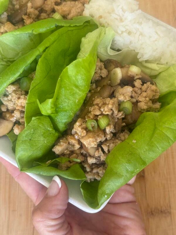 a hand holding a white dish full of chicken lettuce wraps and white rice