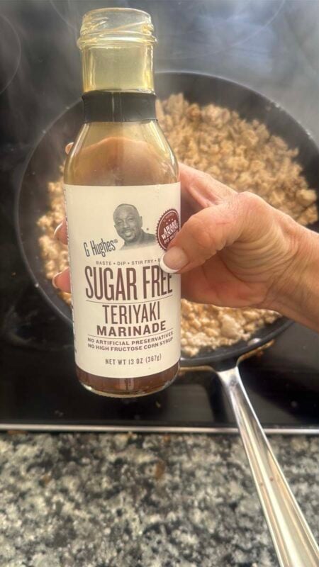 G Hughes sugar free teriyaki marinade being held up over a pan of ground chicken cooking on a stovetop