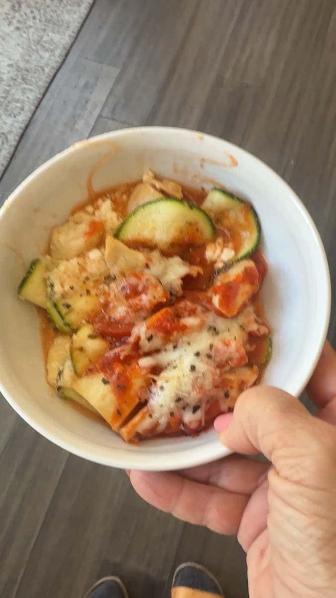 hand holding a white bowl with a chicken parmesan zucchini mixture in it